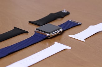A smartwatch with a blue band lies on a wooden surface, surrounded by interchangeable wristbands in black and white colors. The watch has a metallic frame, and the display is turned off. The arrangement is neatly organized, indicating a focus on accessories.
