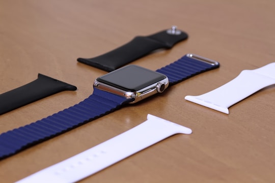 A smartwatch with a blue band lies on a wooden surface, surrounded by interchangeable wristbands in black and white colors. The watch has a metallic frame, and the display is turned off. The arrangement is neatly organized, indicating a focus on accessories.
