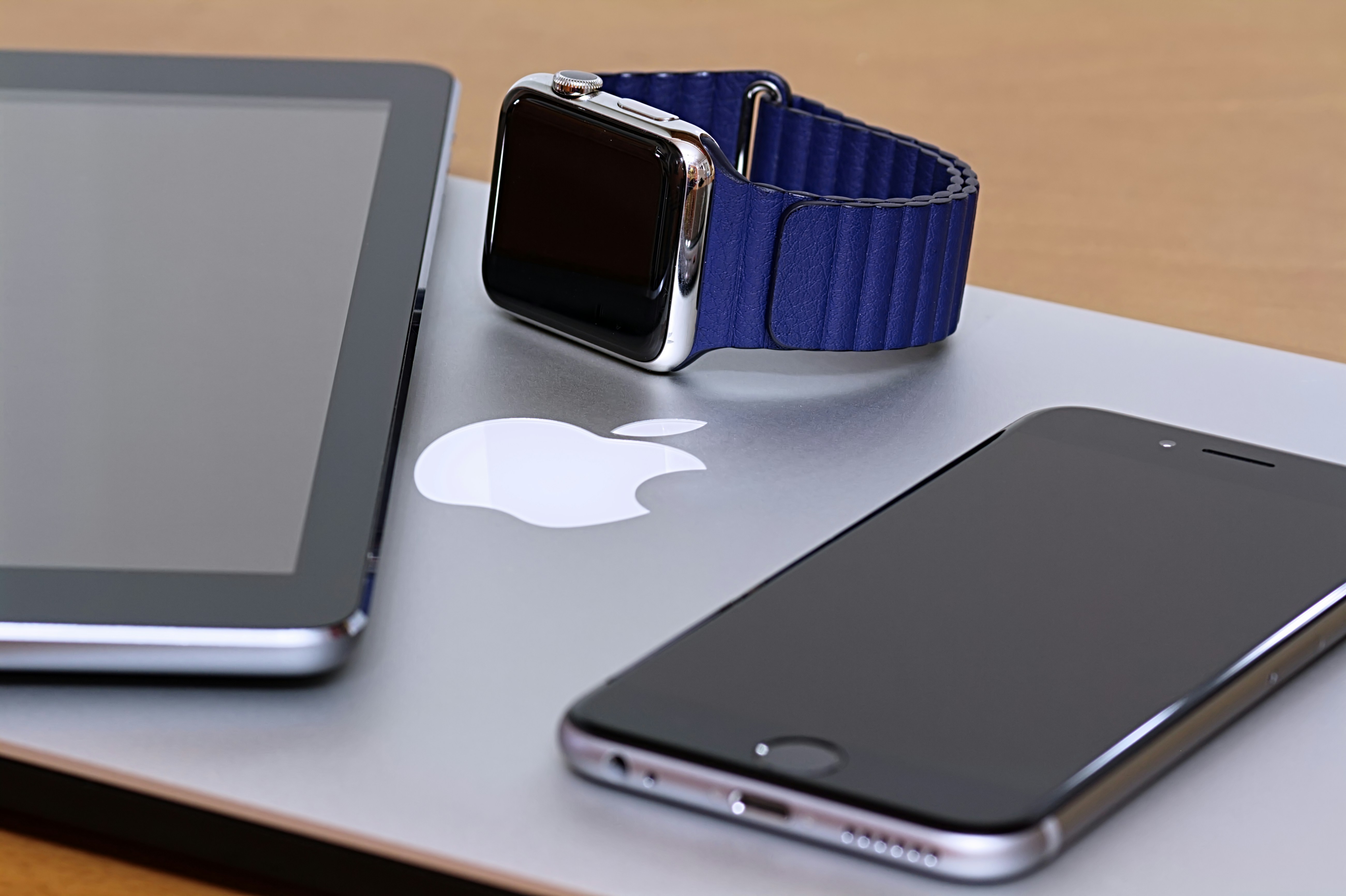 A collection of modern gadgets is arranged on a wooden surface, including a tablet, a smartwatch with a blue band, and a smartphone, all placed on top of a laptop with a visible Apple logo.