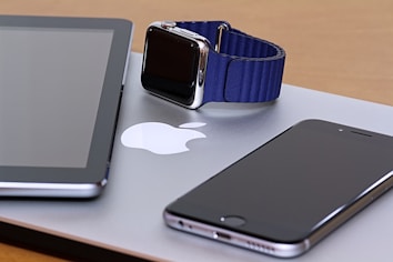 A collection of modern gadgets is arranged on a wooden surface, including a tablet, a smartwatch with a blue band, and a smartphone, all placed on top of a laptop with a visible Apple logo.