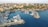 An aerial view of a coastal city with a marina extending into a calm blue sea. The area is populated with numerous boats docked at the piers and surrounded by historic buildings. Lush greenery intersperses the cityscape, and a lighthouse is visible at the end of the breakwater. The city is bordered by natural rocky formations along the coastline.