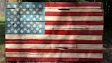 A rustic, wooden representation of the American flag with blue, white, and red painted slats and star patterns. The wood appears weathered and worn, giving it a vintage appearance.