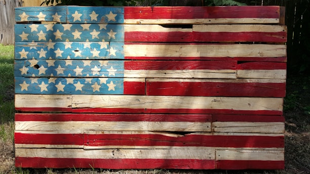 A rustic, wooden representation of the American flag with blue, white, and red painted slats and star patterns. The wood appears weathered and worn, giving it a vintage appearance.