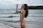 A woman in a colorful swimsuit is standing on a beach with waves in the background. She holds a clear bottle up to her mouth to drink, with her long hair cascading down her back. The sky is cloudy and the ocean appears to be calm.