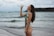 A woman in a colorful swimsuit is standing on a beach with waves in the background. She holds a clear bottle up to her mouth to drink, with her long hair cascading down her back. The sky is cloudy and the ocean appears to be calm.