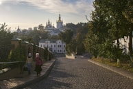 A cobblestone pathway leads up to a grand religious complex with golden domes, surrounded by lush greenery and trees. Two figures are walking along the path, wearing headscarves, and the scene is bathed in morning light creating a serene atmosphere.