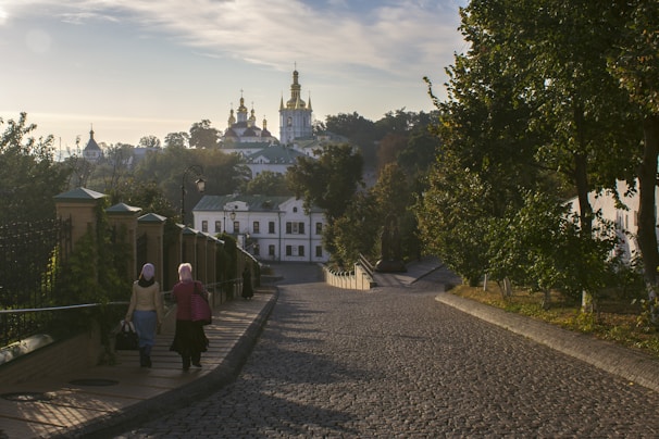 A cobblestone pathway leads up to a grand religious complex with golden domes, surrounded by lush greenery and trees. Two figures are walking along the path, wearing headscarves, and the scene is bathed in morning light creating a serene atmosphere.