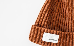 A close-up view of a knitted brown beanie with a folded brim. Attached to the brim is a white label displaying the word 'Hopeless' in black letters. The background is a plain white.