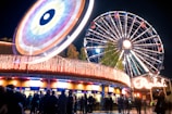 A night scene at an amusement park featuring a brightly lit Ferris wheel and a spinning ride, with colorful lights creating vibrant patterns. A crowd of people is gathered near a ticket counter, and there are trees visible in the background.