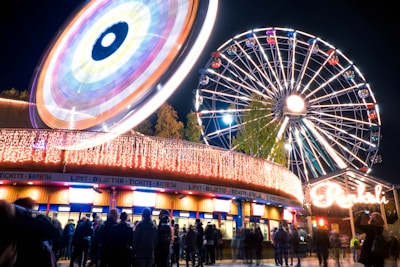 A night scene at an amusement park featuring a brightly lit Ferris wheel and a spinning ride, with colorful lights creating vibrant patterns. A crowd of people is gathered near a ticket counter, and there are trees visible in the background.