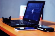 A wooden desk holds an open black laptop displaying a dark screen with a subtle design. Next to the laptop, there is a black lens hood, a smartphone with a reflective screen, and a portable gaming console. The background is out of focus, showcasing a blurred calendar and a neutral-colored wall.