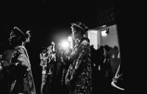 A group of people wearing traditional attire stand in a dimly lit area. One person is illuminated by light, suggesting a focus or spotlight. The background appears to have photographers or onlookers, some holding cameras. The setting gives a sense of anticipation or preparation, possibly before or after an event.