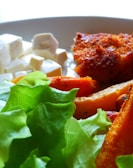 A close-up of a plate containing fresh green lettuce, white cubed cheese, crispy roasted sweet potatoes, and a piece of breaded meat. The textures and colors of the food items are vivid, with light softly illuminating the scene.