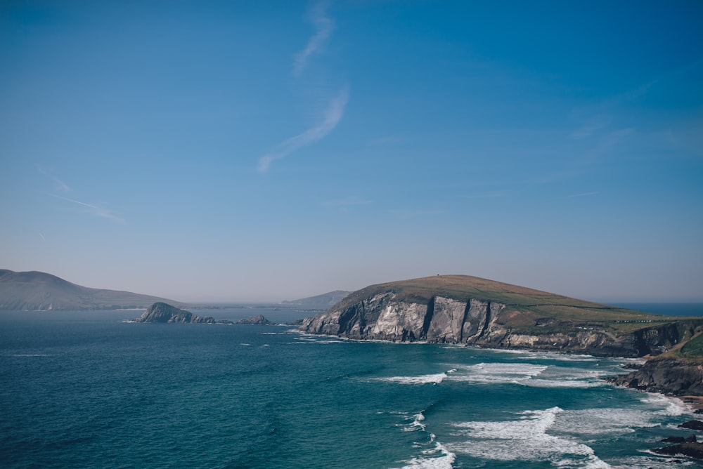 ondas do mar ao lado de penhasco cinza e verde sob céu azul claro durante o dia