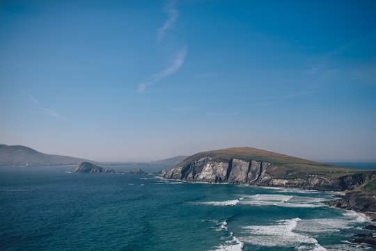 sea waves beside gray and green cliff under clear blue sky during daytime in Dingle Peninsula Ireland