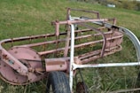 An old and rusted piece of agricultural machinery lies on a grassy field. The metal components are primarily pink, with visible rust and wear. It has intricate details with loops and rotating mechanisms, suggesting it may have been used for harvesting or processing crops in the past.