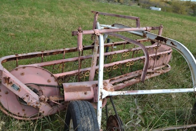 An old and rusted piece of agricultural machinery lies on a grassy field. The metal components are primarily pink, with visible rust and wear. It has intricate details with loops and rotating mechanisms, suggesting it may have been used for harvesting or processing crops in the past.
