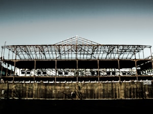 A rusted, skeletal structure of a large industrial building stands against a clear sky. The exposed metal framework is expansive, with shadows creating intricate patterns across the remaining walls. The lower section appears to be made of concrete, showing signs of wear and graffiti. Sparse vegetation can be seen in the background, adding a hint of nature reclaiming the space.