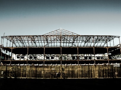 A rusted, skeletal structure of a large industrial building stands against a clear sky. The exposed metal framework is expansive, with shadows creating intricate patterns across the remaining walls. The lower section appears to be made of concrete, showing signs of wear and graffiti. Sparse vegetation can be seen in the background, adding a hint of nature reclaiming the space.