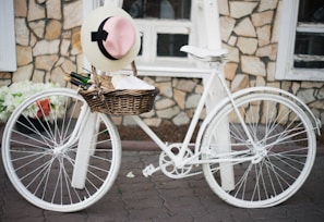 A vintage-style white bicycle with a wicker basket attached to the front, resting against a textured stone wall. The basket contains a straw hat with a pink ribbon, a bottle, and some rolled garments. The setting gives a rustic and charming ambiance.