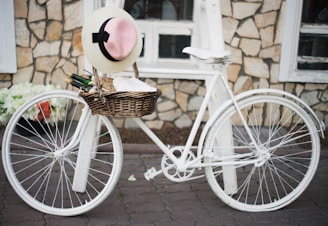 A vintage-style white bicycle with a wicker basket attached to the front, resting against a textured stone wall. The basket contains a straw hat with a pink ribbon, a bottle, and some rolled garments. The setting gives a rustic and charming ambiance.