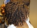 A person with tightly coiled, shiny hair is being styled. The hair has a springy texture, and a hand is visible holding scissors, suggesting a haircut or styling in progress. The setting appears to be indoors, possibly a salon.