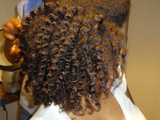 A person with tightly coiled, shiny hair is being styled. The hair has a springy texture, and a hand is visible holding scissors, suggesting a haircut or styling in progress. The setting appears to be indoors, possibly a salon.