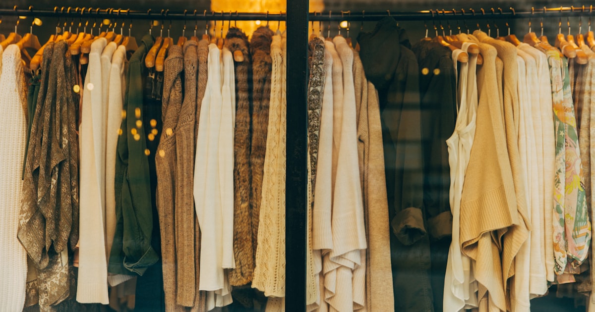 Two clothing racks displayed in a warm-lit boutique window filled with earth-tone and neutral fashion pieces