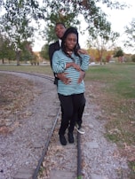 A couple is standing on a set of narrow train tracks in a park setting surrounded by green and autumn-colored trees. The woman is wearing a striped shirt and leggings, while the man is in a dark jacket and standing closely behind her. They appear relaxed and affectionate.