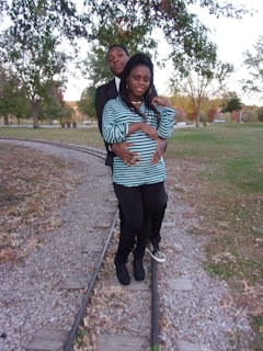 A couple is standing on a set of narrow train tracks in a park setting surrounded by green and autumn-colored trees. The woman is wearing a striped shirt and leggings, while the man is in a dark jacket and standing closely behind her. They appear relaxed and affectionate.