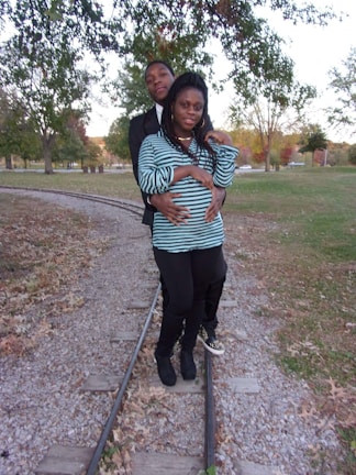 A couple is standing on a set of narrow train tracks in a park setting surrounded by green and autumn-colored trees. The woman is wearing a striped shirt and leggings, while the man is in a dark jacket and standing closely behind her. They appear relaxed and affectionate.