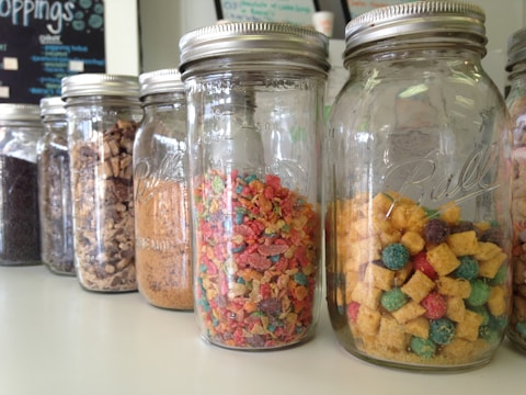 Several glass jars are arranged in a line on a countertop, each filled with different types of colorful cereals and dry food items. The jars have metal lids, and the labels on the jars read 'Ball'. In the background, part of a menu or sign is partially visible.