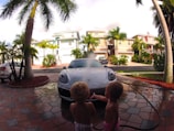 Two young children are playing near a sports car that is being washed with water. The scene is set on a palm-lined driveway with several pastel-colored houses in the background. The driveway is made of decorative stone tiles, and a hose is visible, spraying water over the car.