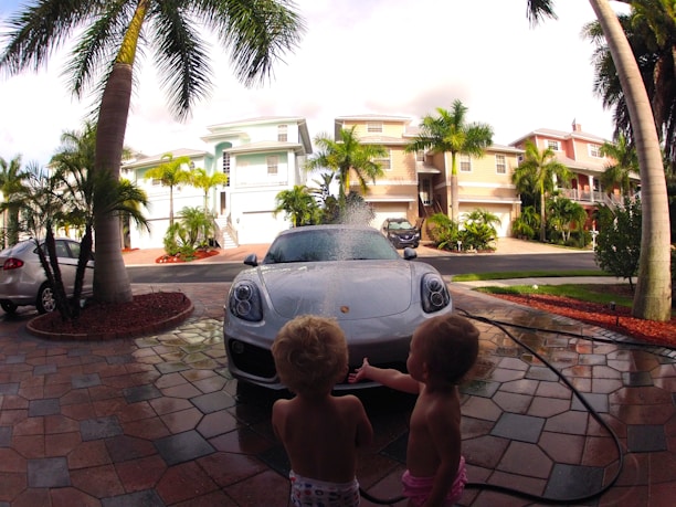 Two young children are playing near a sports car that is being washed with water. The scene is set on a palm-lined driveway with several pastel-colored houses in the background. The driveway is made of decorative stone tiles, and a hose is visible, spraying water over the car.