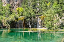 A serene natural landscape featuring a small waterfall cascading over rocky cliffs into a clear, turquoise pool. Surrounding the water are dense, lush green trees and vegetation, with the rock face adding texture and depth to the scene.
