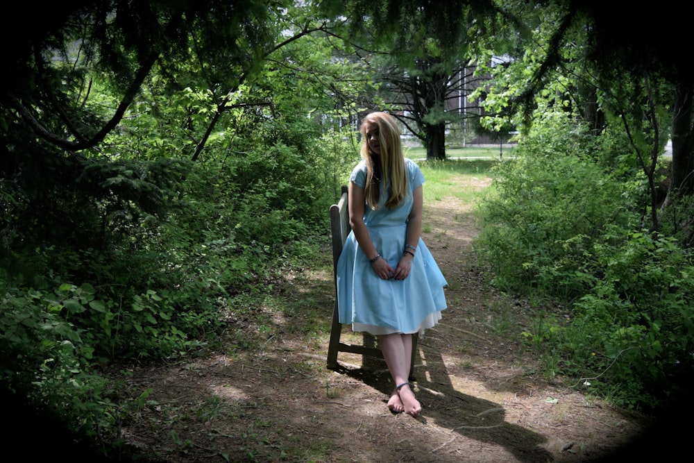 A young adult female in a white dress leaning on a chair in the middle of the woods.