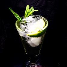 A tall glass filled with a refreshing beverage, featuring ice cubes, lime slices, and fresh mint leaves. The background is dark, highlighting the vibrant green and translucent colors of the drink.