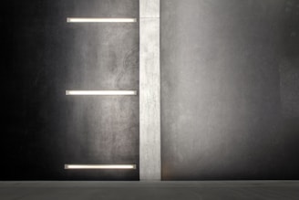 A modern, minimalist wall design with a vertical light gray panel separating two dark gray sections. Horizontal fluorescent-style lights are embedded on the left section of the wall, providing a muted illumination.