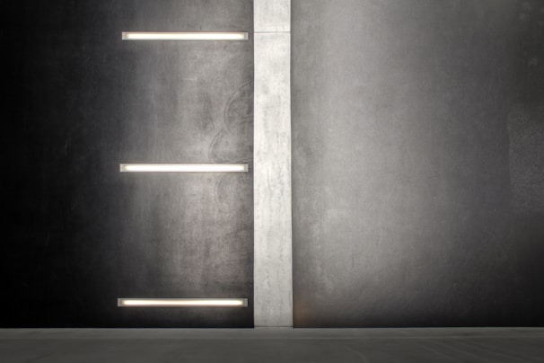 A modern, minimalist wall design with a vertical light gray panel separating two dark gray sections. Horizontal fluorescent-style lights are embedded on the left section of the wall, providing a muted illumination.