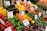 A vibrant and colorful display of fresh produce at a market stall. Various fruits and vegetables are neatly arranged with noticeable price and product signs. Items include green, red, and yellow bell peppers, eggplant, lemons, apples, bananas, tomatoes, and baby potatoes, among others. The setup creates a lively and inviting atmosphere.