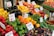 A vibrant and colorful display of fresh produce at a market stall. Various fruits and vegetables are neatly arranged with noticeable price and product signs. Items include green, red, and yellow bell peppers, eggplant, lemons, apples, bananas, tomatoes, and baby potatoes, among others. The setup creates a lively and inviting atmosphere.