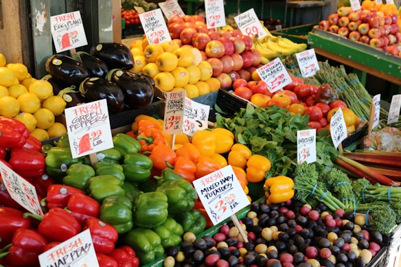 A vibrant and colorful display of fresh produce at a market stall. Various fruits and vegetables are neatly arranged with noticeable price and product signs. Items include green, red, and yellow bell peppers, eggplant, lemons, apples, bananas, tomatoes, and baby potatoes, among others. The setup creates a lively and inviting atmosphere.
