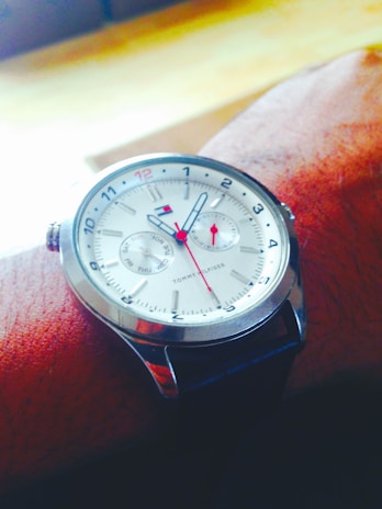 A wristwatch with a silver casing and a white watch face, featuring blue and red accents. It has a leather strap and displays detailed watch hands with additional small dials for day tracking.