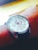 A wristwatch with a silver casing and a white watch face, featuring blue and red accents. It has a leather strap and displays detailed watch hands with additional small dials for day tracking.