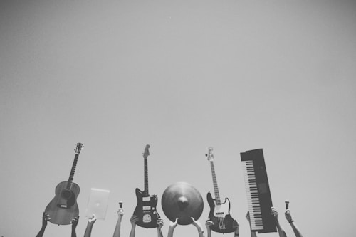 Various musical instruments and objects are being held up against a clear sky. Visible items include an acoustic guitar, an electric guitar, a bass guitar, a cymbal, a keyboard, and a laptop.