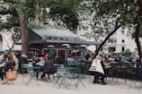 A bustling outdoor setting filled with people enjoying food and drinks at green metal tables and chairs. In the background, there is a food kiosk with greenery and a large sign on top. Surrounding the area are tall trees and string lights, creating a relaxed, open-air atmosphere.