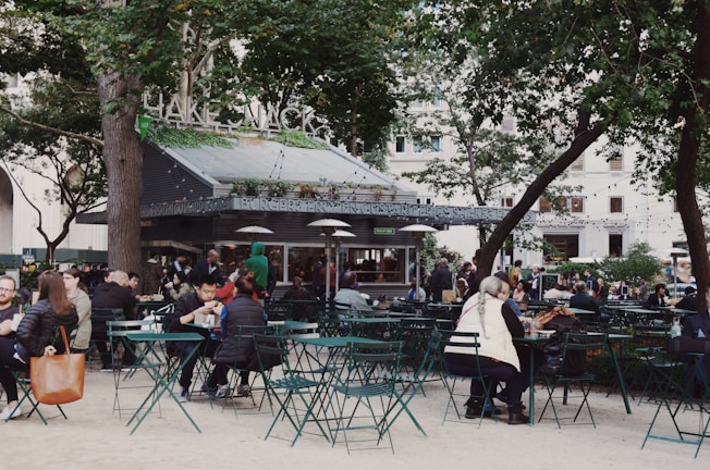 A bustling outdoor setting filled with people enjoying food and drinks at green metal tables and chairs. In the background, there is a food kiosk with greenery and a large sign on top. Surrounding the area are tall trees and string lights, creating a relaxed, open-air atmosphere.