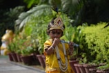 A child is dressed in traditional attire resembling a deity with a colorful headdress and a beaded necklace, playing a flute amidst a garden setting with lush green foliage.