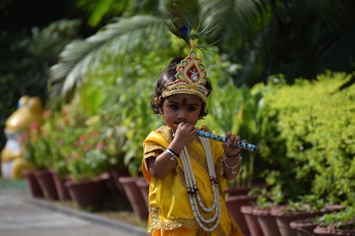 A child is dressed in traditional attire resembling a deity with a colorful headdress and a beaded necklace, playing a flute amidst a garden setting with lush green foliage.