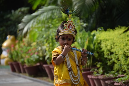 A child is dressed in traditional attire resembling a deity with a colorful headdress and a beaded necklace, playing a flute amidst a garden setting with lush green foliage.